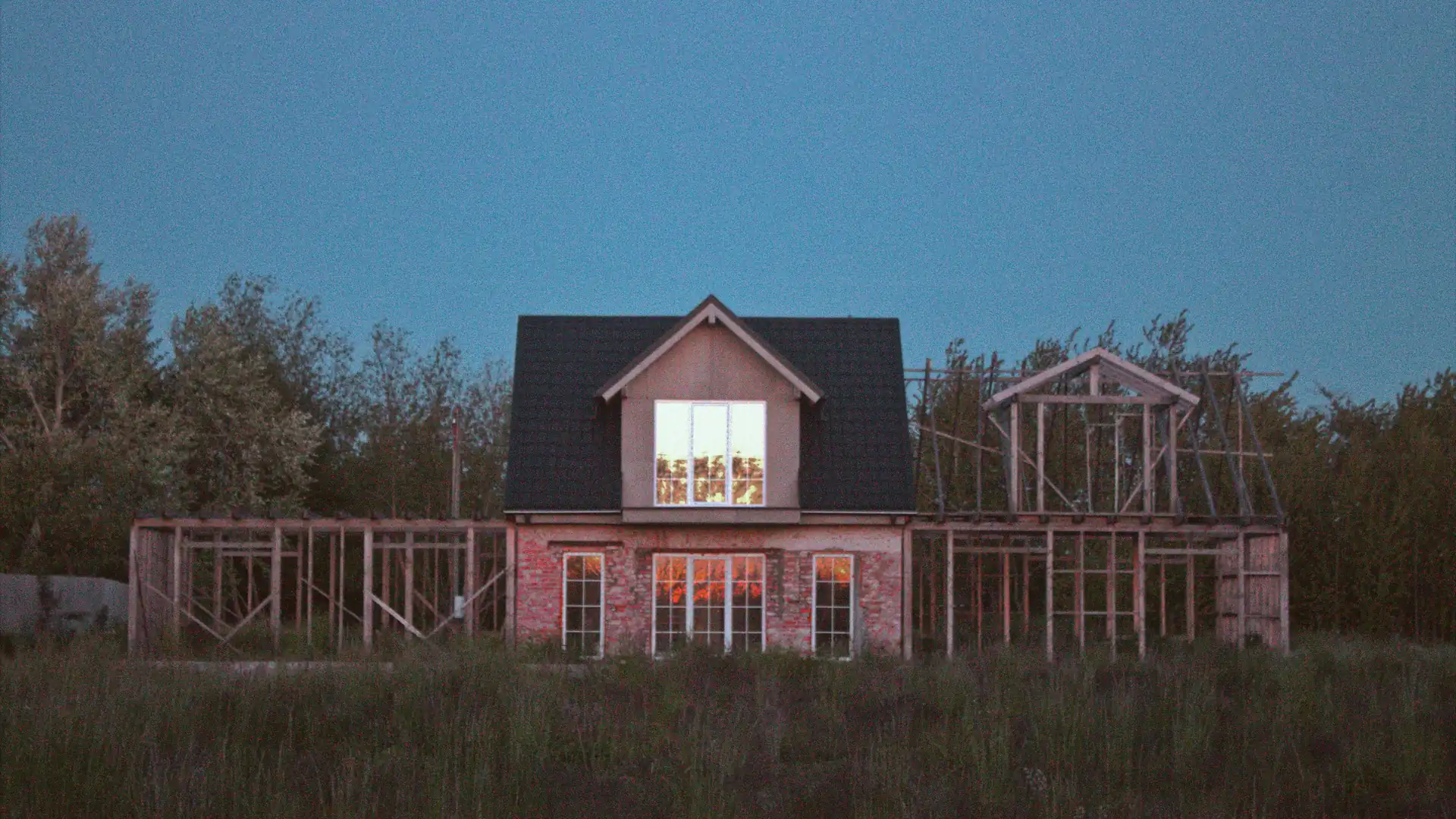 Halbfertiges Haus im Feld zur blauen Stunde, die eingebauten Fenster leuchten warm aus dem Rohbau.