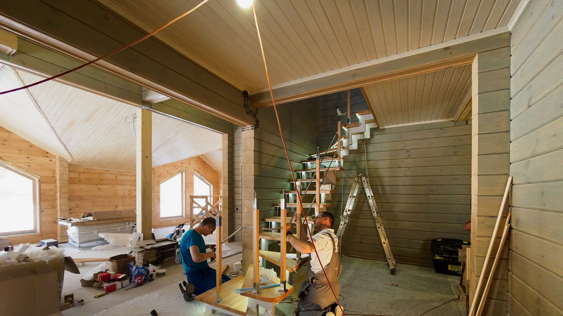 Zwei Handwerker montieren eine Holztreppe mit Mittelholm in einem noch unfertigen Holzhaus, darueber eine einzelne Gluehbirne.